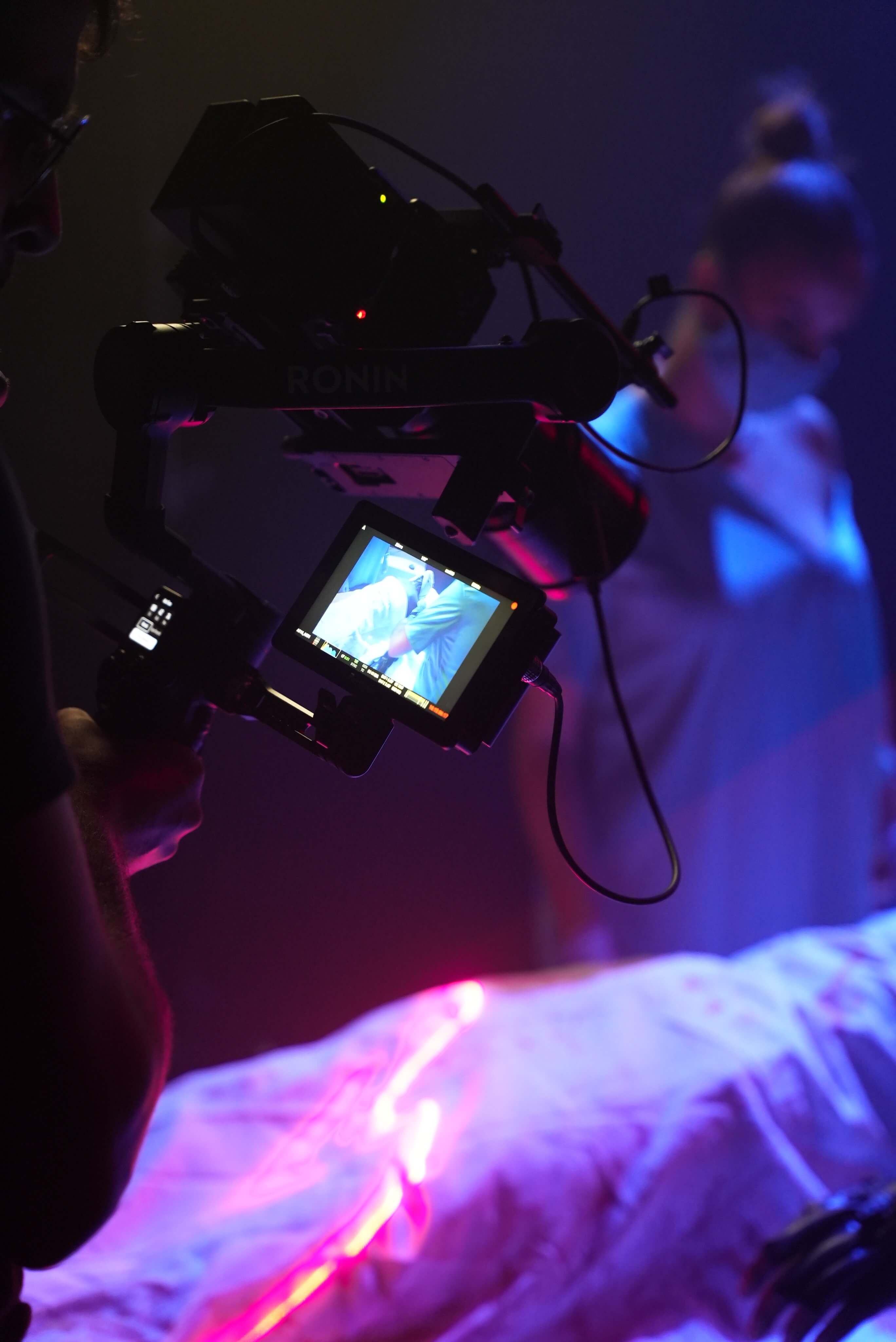 A music video shooting with sci fi lighting, the videographer holding a DJ Rs3 pro stabilzer with a Red Komodo 6k paird with DZOFILM 20-55mm T2.8 Pictor Zoom on the top, a nurse visible in the background and some white sheet.