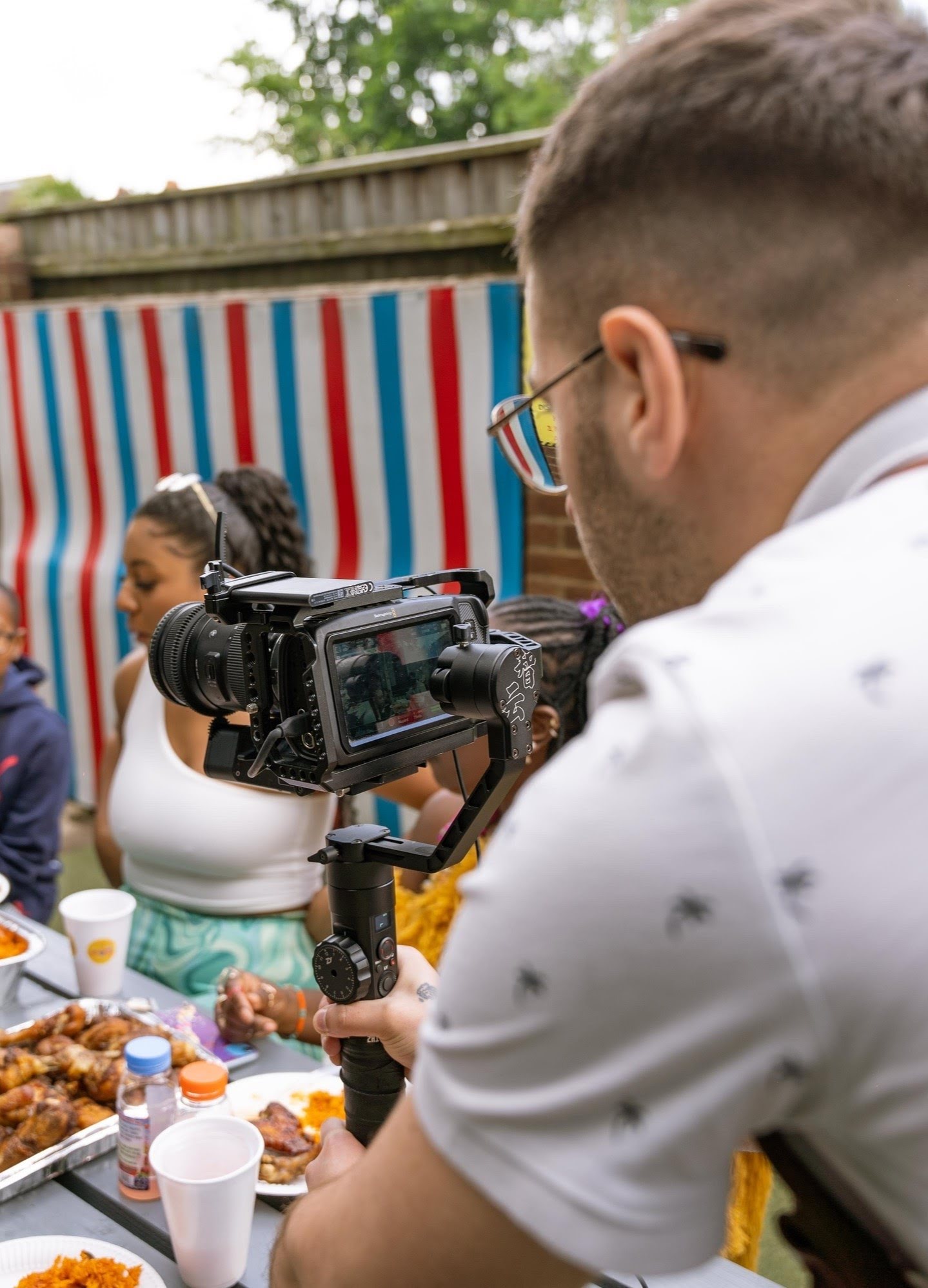 Videographer shooting with a Black Magick Pocket (BMPCC) 4K and a Zhiyun Carne 2 stabilzer for msuic video at the table with a lots of food and some girls in the background.